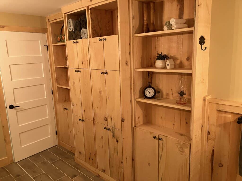 A wooden cabinet with multiple doors and open shelves is against a wall. The shelves hold rolled towels, small plants, a clock, and decorative items. The cabinet is next to a closed white door on a tiled floor.