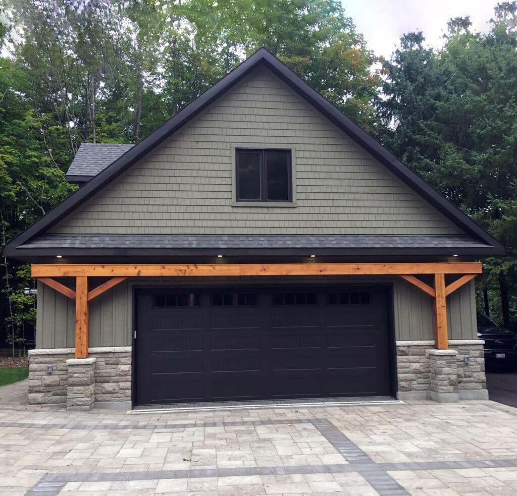 A modern garage with a pitched roof and dark brown double doors is set against a backdrop of trees. The structure features stone and wood accents, with a paved driveway in the foreground.