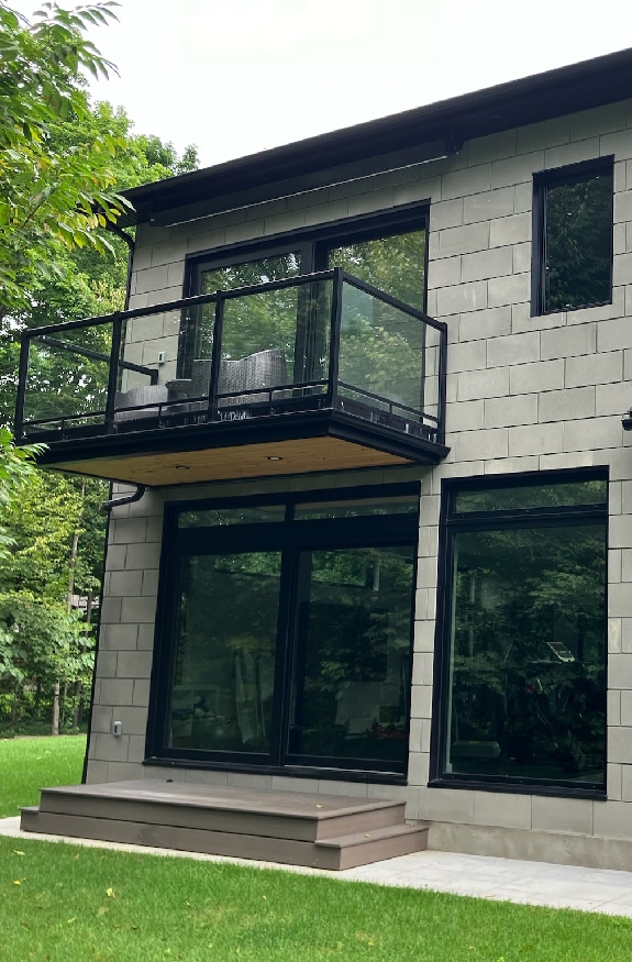 A modern two-story house with a beige brick exterior, black-framed windows, and a glass balcony on the upper level. It is surrounded by green grass and leafy trees.
