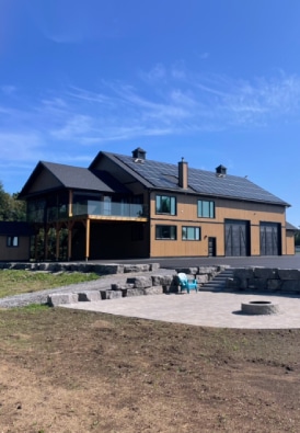 A large modern house with solar panels on the roof. The house has a wooden exterior with large windows and a covered porch. In the foreground, there is a patio area with a fire pit and a person seated in a chair. The sky is clear and blue.